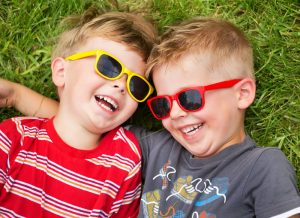 Two toddler boys with colorful sunglasses on laugh on top of artificial turf in New Jersey